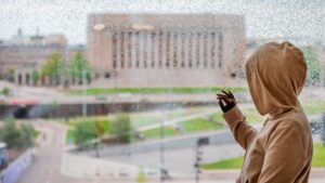 Ihminen katsoo Suomen eduskuntataloa sateisen ikkunaruudun läpi.