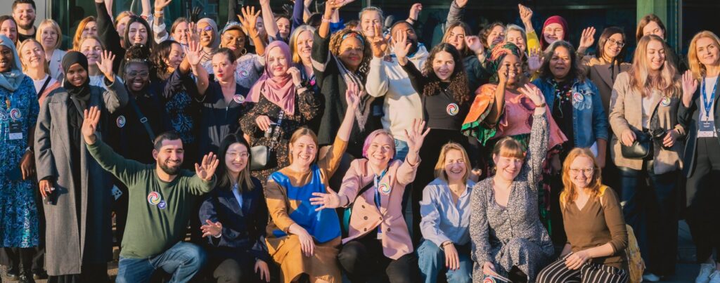 joukko ihmisiä eri taustoista poseeraa yhdessä ja vilkuttaa kameralle